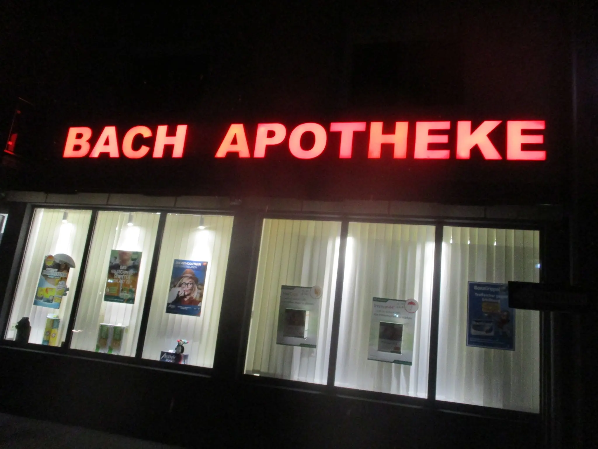 Rot leuchtender Schriftzug „Bach Apotheke“ über beleuchteten Schaufenstern bei Nacht